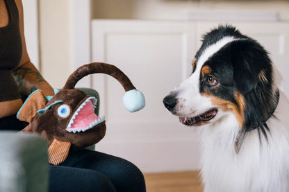 Black and white dog playing with brown angler fish plush toy designed for interactive enrichment